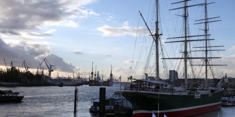 Hinter dem Dreimaster zeigen sich die großen Kräne, die Stückgut verladen.