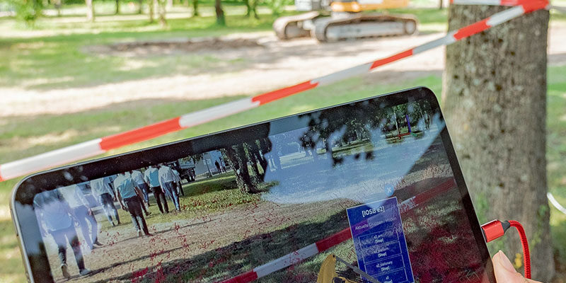 Bagger (Hintergrund) mit digitalem Zwilling auf einem Tablet für Augmented-Reality durch Technologie des KIT-ISAS. Eingeblendet werden Sensordaten des Baggers als gefärbte Punktwolke. Es besteht auch die Möglichkeit zur Fernsteuerung des Baggers durch das Tablet. (Foto: ROBDEKON / Fraunhofer-Institut für Optronik, Systemtechnik und Bildauswertung IOSB)